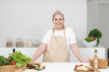 A mature woman in an apron stands at a kitchen counter with fresh produce and cheese, ready for cooking.